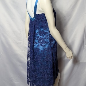 May include: A sleeveless, knee-length dress in shades of blue, displayed on a mannequin. The dress features a dark blue floral lace overlay over a lighter blue fabric. The straps are a darker blue satin. The dress has a loose, flowing silhouette.