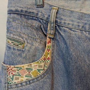 Può includere: Primo piano di jeans di denim blu chiaro con dettagli decorativi sulle tasche. Le tasche presentano un bordo in tessuto colorato a fantasia con disegni floreali e geometrici. I jeans sono appesi a una gruccia di legno.