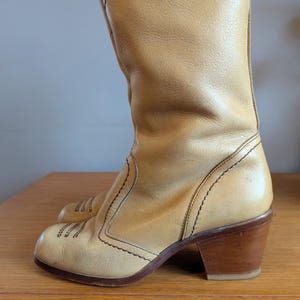 Op de afbeelding: Een paar bruinleren laarzen met een ronde neus en een gestapelde houten hak. De laarzen hebben decoratieve stiksels en een kniehoge schacht. De hak meet ongeveer 6,35 cm.