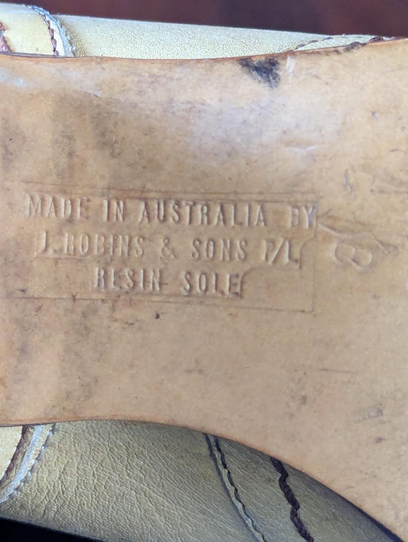 Op de afbeelding: Close-up van een schoenzool met de tekst "MADE IN AUSTRALIA BY J. ROBINS & SONS P/L RESIN SOLE". De zool is lichtbruin van kleur en vertoont slijtage.