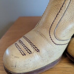 Op de afbeelding: Close-up van een beige leren laars met bruine stiksels en een bruine zool. De laars heeft een puntige neus en een blokhak. Het leer lijkt versleten, met zichtbare kreukels en krassen, wat duidt op een vintage of geliefd item.