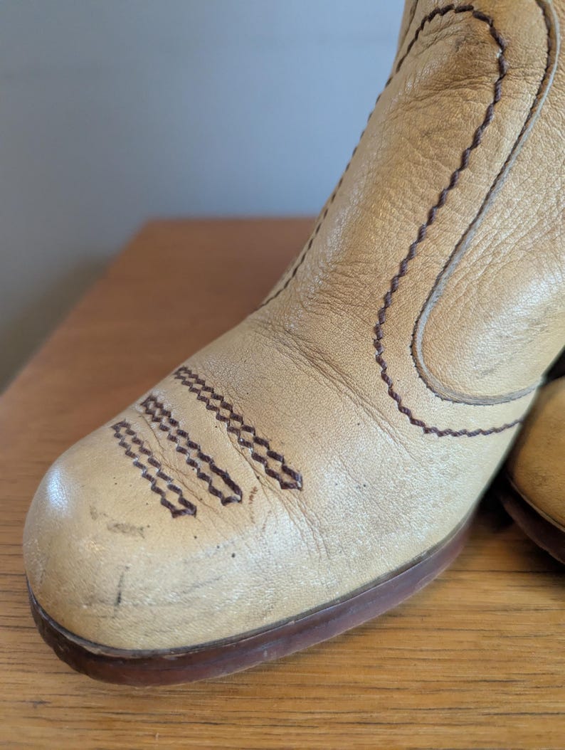 Op de afbeelding: Close-up van een beige leren laars met bruine stiksels en een bruine zool. De laars heeft een puntige neus en een blokhak. Het leer lijkt versleten, met zichtbare kreukels en krassen, wat duidt op een vintage of geliefd item.