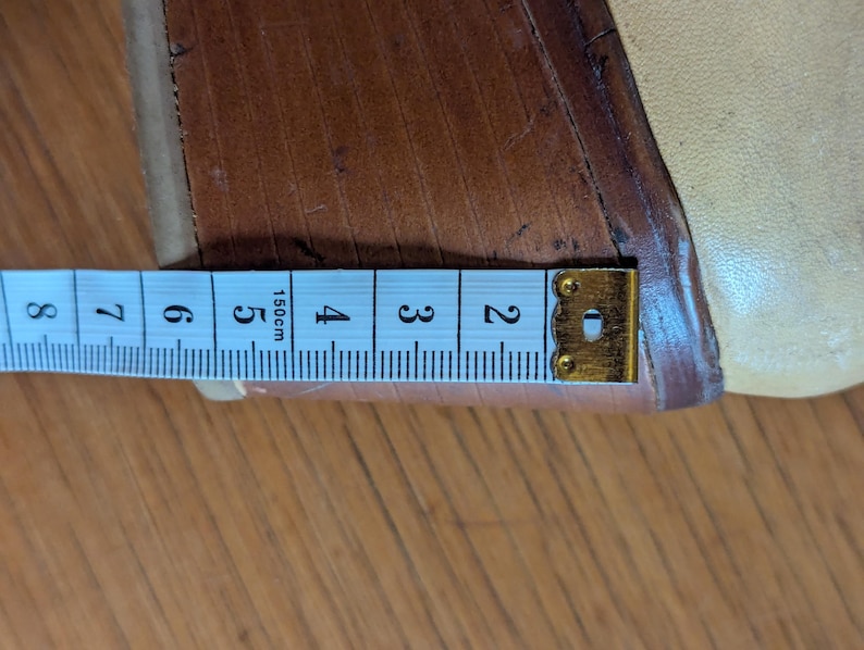 Op de afbeelding: Een close-up toont een beige schoen met een houten sleehak. Een meetlint is naast de hak geplaatst, met een meting van ongeveer 5 cm.