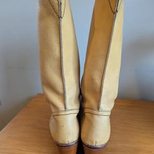 Op de afbeelding: Een paar hoge cowboylaarzen van lichtbruin leer. De laarzen hebben een puntige bovenkant en een bruine houten hak. De stiksels zijn zichtbaar en de laarzen lijken vintage te zijn.