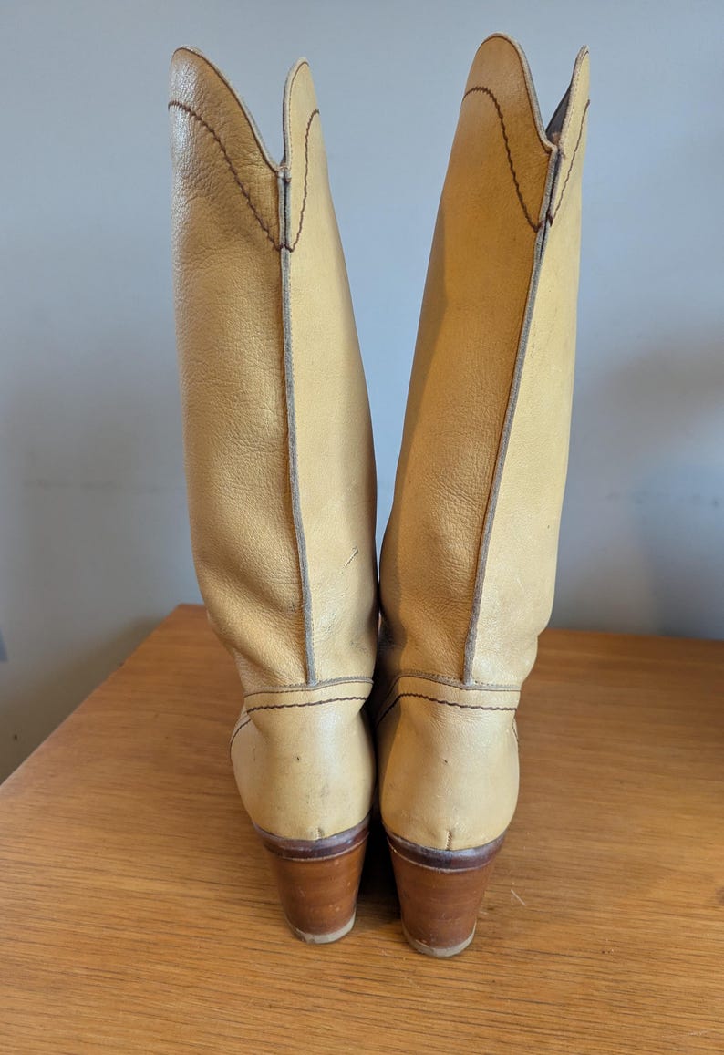 Op de afbeelding: Een paar hoge cowboylaarzen van lichtbruin leer. De laarzen hebben een puntige bovenkant en een bruine houten hak. De stiksels zijn zichtbaar en de laarzen lijken vintage te zijn.