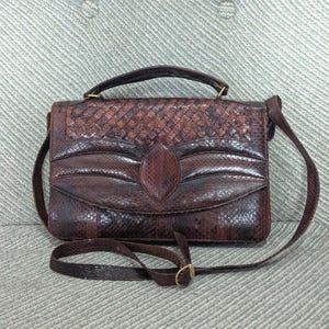 May include: A brown leather handbag with a woven top flap and a decorative leaf-shaped accent. The bag has a top handle and a long, adjustable shoulder strap. The hardware is gold-toned.