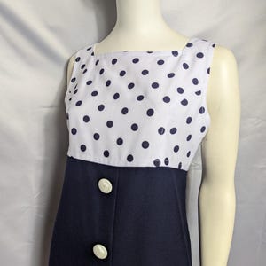 May include: A sleeveless dress featuring a white bodice with navy blue polka dots and a solid navy blue skirt section. The front of the dress includes a vertical row of three large, round, off-white buttons positioned along the centre seam.