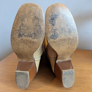 Op de afbeelding: Een paar vintage beige leren laarzen, maat 37, met houten blokhakken. De zolen vertonen slijtage. De laarzen staan op een houten oppervlak.