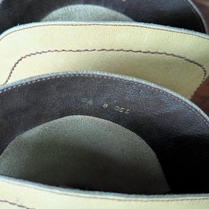 Op de afbeelding: Close-up van de binnenkant van drie leren laarzen. De laarzen zijn in tinten beige en bruin. De stiksels zijn zichtbaar langs de randen. De nummers "9 8 055" zijn in de laarzen gestempeld.