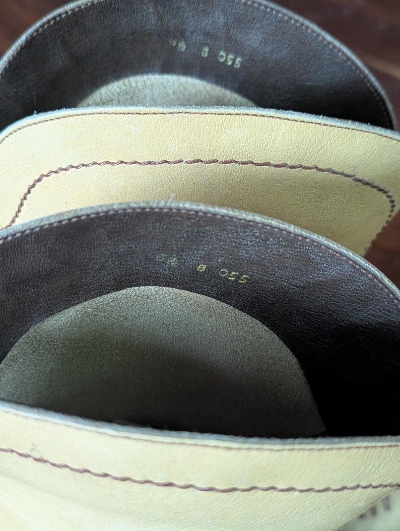 Op de afbeelding: Close-up van de binnenkant van drie leren laarzen. De laarzen zijn in tinten beige en bruin. De stiksels zijn zichtbaar langs de randen. De nummers "9 8 055" zijn in de laarzen gestempeld.