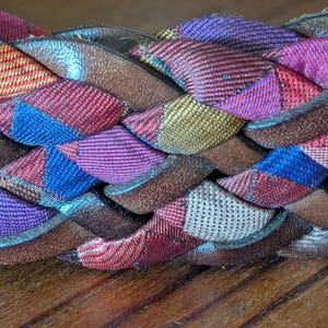 Può includere: Primo piano di una cintura intrecciata. La cintura presenta un motivo tessuto con sezioni di tessuto rosso, blu, viola e oro. Il rivestimento in pelle è marrone. La cintura è appoggiata su una superficie di legno.