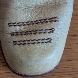 Op de afbeelding: Close-up van een leren laarsneus in beige. De laars heeft decoratieve bruine stiksels langs de bovenkant en over de neus. Het leer heeft een versleten uiterlijk.
