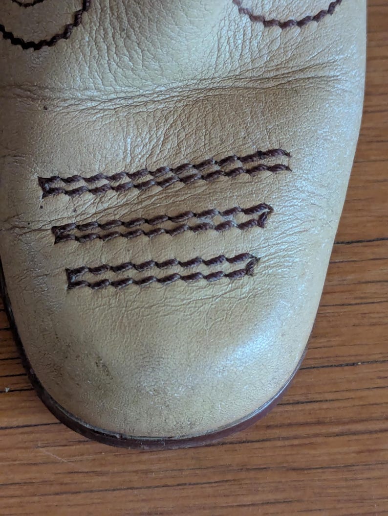 Op de afbeelding: Close-up van een leren laarsneus in beige. De laars heeft decoratieve bruine stiksels langs de bovenkant en over de neus. Het leer heeft een versleten uiterlijk.