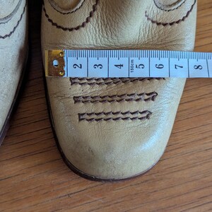 Op de afbeelding: Close-up van een paar lichtbruine leren laarzen met decoratieve stiksels. Een meetlint toont de breedte van de laars. De laarzen hebben een Western-stijl met geschulpte randen en gedetailleerde stiksels.