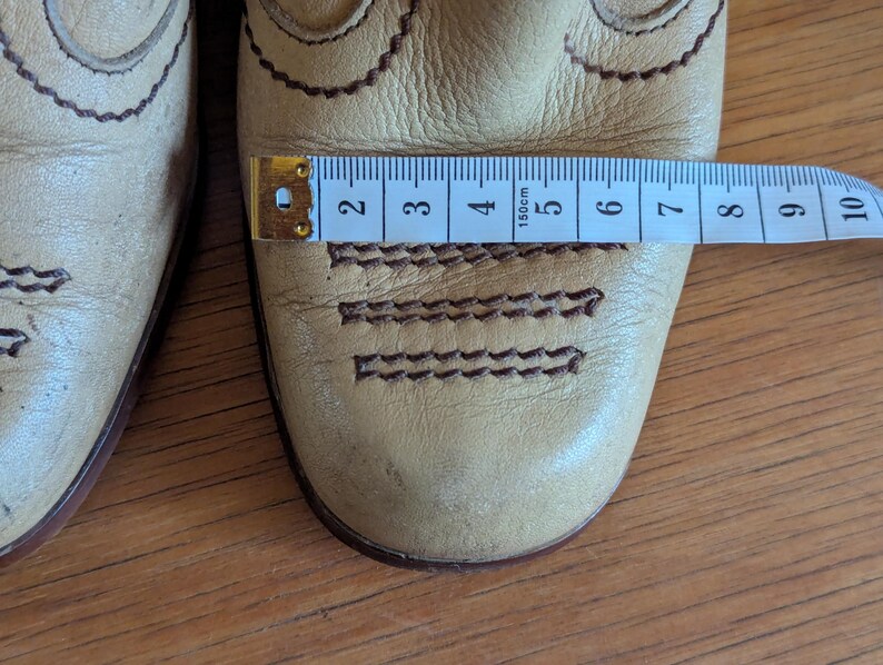 Op de afbeelding: Close-up van een paar lichtbruine leren laarzen met decoratieve stiksels. Een meetlint toont de breedte van de laars. De laarzen hebben een Western-stijl met geschulpte randen en gedetailleerde stiksels.