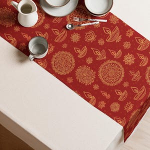 May include: A red table runner with a repeating gold pattern of mandalas, lotus flowers, and oil lamps. The runner is on a white table, with a white pitcher, a mug, and tableware visible.