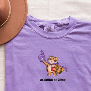 A purple short-sleeved t-shirt with a cartoon tiger wearing a purple cape and a foam finger, holding a red cup, with the text "WE DRINK AT DAWN" below it.