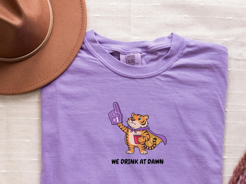 A purple short-sleeved t-shirt with a cartoon tiger wearing a purple cape and a foam finger, holding a red cup, with the text "WE DRINK AT DAWN" below it.