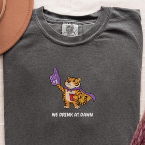 A dark gray short-sleeved t-shirt with a cartoon tiger wearing a purple cape and a foam finger, holding a red cup, with the text "WE DRINK AT DAWN" below it.