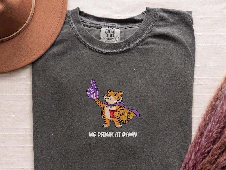 A dark gray short-sleeved t-shirt with a cartoon tiger wearing a purple cape and a foam finger, holding a red cup, with the text "WE DRINK AT DAWN" below it.