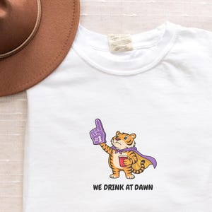 A white short-sleeved t-shirt with a cartoon tiger wearing a purple cape and a foam finger, holding a red cup, with the text "WE DRINK AT DAWN" below it.