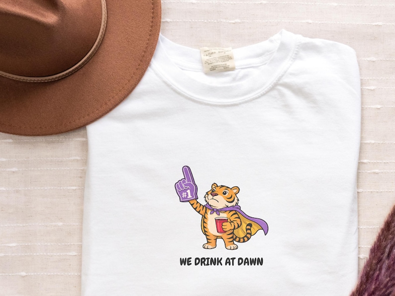 A white short-sleeved t-shirt with a cartoon tiger wearing a purple cape and a foam finger, holding a red cup, with the text "WE DRINK AT DAWN" below it.