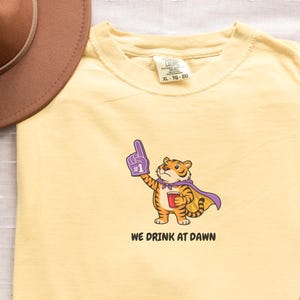 A light yellow short-sleeved t-shirt with a cartoon tiger wearing a purple cape and a foam finger, holding a red cup, with the text "WE DRINK AT DAWN" below it.