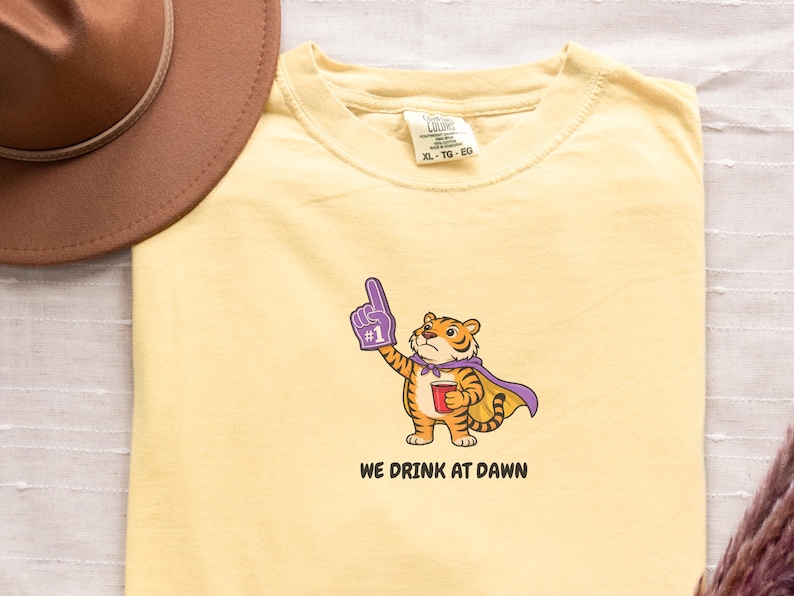 A light yellow short-sleeved t-shirt with a cartoon tiger wearing a purple cape and a foam finger, holding a red cup, with the text "WE DRINK AT DAWN" below it.