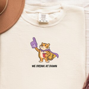 A cream-colored short-sleeved t-shirt with a cartoon tiger wearing a purple cape and a foam finger, holding a red cup, with the text "WE DRINK AT DAWN" below it.