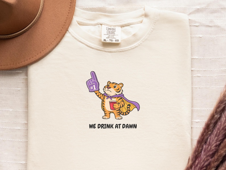 A cream-colored short-sleeved t-shirt with a cartoon tiger wearing a purple cape and a foam finger, holding a red cup, with the text "WE DRINK AT DAWN" below it.