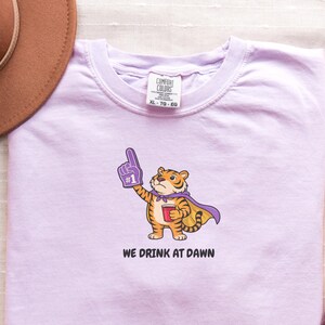 A light purple short-sleeved t-shirt with a cartoon tiger wearing a purple cape and a foam finger, holding a red cup, with the text "WE DRINK AT DAWN" below it.