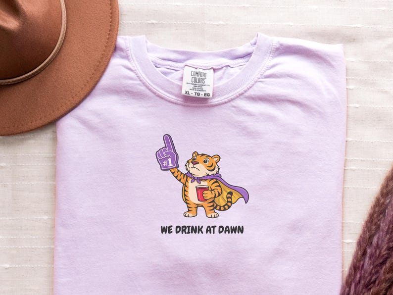 A light purple short-sleeved t-shirt with a cartoon tiger wearing a purple cape and a foam finger, holding a red cup, with the text "WE DRINK AT DAWN" below it.