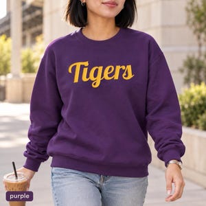 May include: A deep purple crewneck sweatshirt featuring the word Tigers printed in a bold, yellow cursive script across the chest. The garment has long sleeves with ribbed cuffs and a ribbed neckline, providing a standard fit for casual wear.