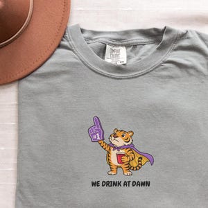 A light gray short-sleeved t-shirt with a cartoon tiger wearing a purple cape and a foam finger, holding a red cup, with the text "WE DRINK AT DAWN" below it.