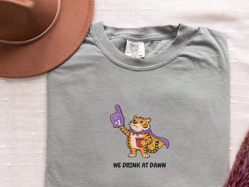 A light gray short-sleeved t-shirt with a cartoon tiger wearing a purple cape and a foam finger, holding a red cup, with the text "WE DRINK AT DAWN" below it.