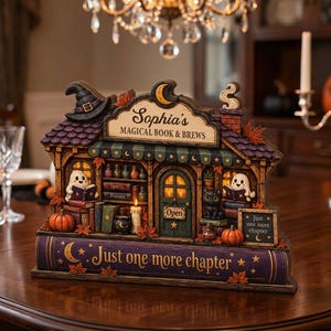 Könnte beinhalten: Eine dekorative Tischdekoration in Form eines Buchladens mit der Aufschrift Sophia's Magical Book & Brews. Das Objekt hat ein violettes Dach, einen Hexenhut, zwei lesende Geister, eine schwarze Katze und einen Sockel wie ein Buchrücken mit dem Text Just one more chapter.
