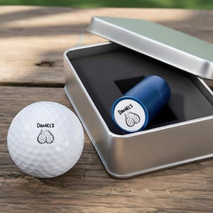 Op de afbeelding: Een witte golfbal en een blauw cilindrisch item met de tekst "DANIEL'S" en een afbeelding van testikels. De items bevinden zich in een zilveren metalen doos met een zwart interieur, op een houten oppervlak.