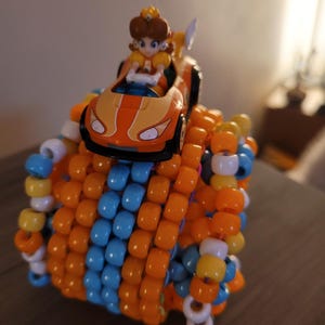 May include: A handmade beaded cuff bracelet in orange, blue, white, and yellow. A small orange race car with a cartoon character figure sits atop the bracelet. The bracelet is made of small, colorful beads.