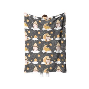 May include: A dark grey blanket featuring a repeating pattern of gnomes, moons, clouds, and stars in tan and yellow. The blanket is held up, displaying the full design.