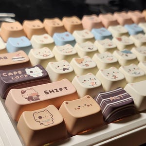 May include: A close-up of a mechanical keyboard with custom keycaps. The keycaps are in pastel shades of peach, cream, and blue. Many keycaps have cat illustrations. The "SHIFT" keycap has a cat graphic.