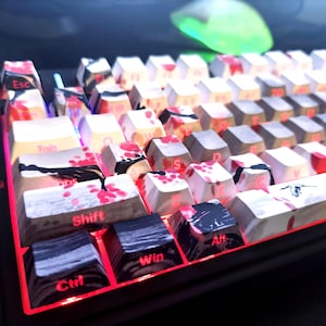 May include: A mechanical keyboard with custom keycaps. The keycaps have a white base with red and black accents. The keyboard has red LED lighting around the keys. The mouse in the background has a green glow.