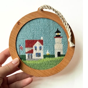 May include: A round, wooden framed cross-stitch featuring a lighthouse and a house with a red roof. The artwork has a blue background and green grass. A white rope is attached for hanging. The frame is held by a hand.