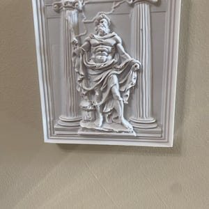 May include: A white relief sculpture of a classical figure, possibly Zeus, holding a staff, framed by two columns. The sculpture is mounted on a square, white frame, and is set against a neutral-toned wall.