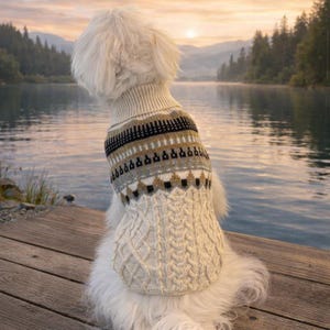 Peut inclure: Un chien blanc porte un pull pour chien en tricot torsadé crème avec un empiècement à motifs. L'empiècement présente des motifs géométriques noirs, dorés et gris. Le chien est assis sur un ponton en bois, surplombant un lac au coucher du soleil.