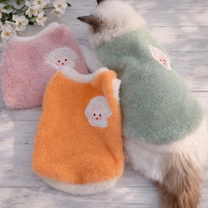 Op de afbeelding: Drie huisdier truien in roze, oranje en groen. Elke trui heeft een witte rand en een cartoon karakter patch. De truien zijn gemaakt van een zacht, pluizig materiaal. De kat draagt een groene trui.