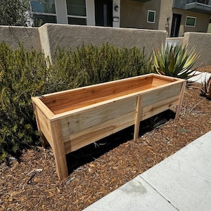 Puede incluir: Una jardinera rectangular de madera con un acabado natural en color marr&oacute;n claro, que cuenta con cuatro patas resistentes y un interior profundo y abierto. La jardinera est&aacute; construida con tablones de madera horizontales y un borde superior reforzado.