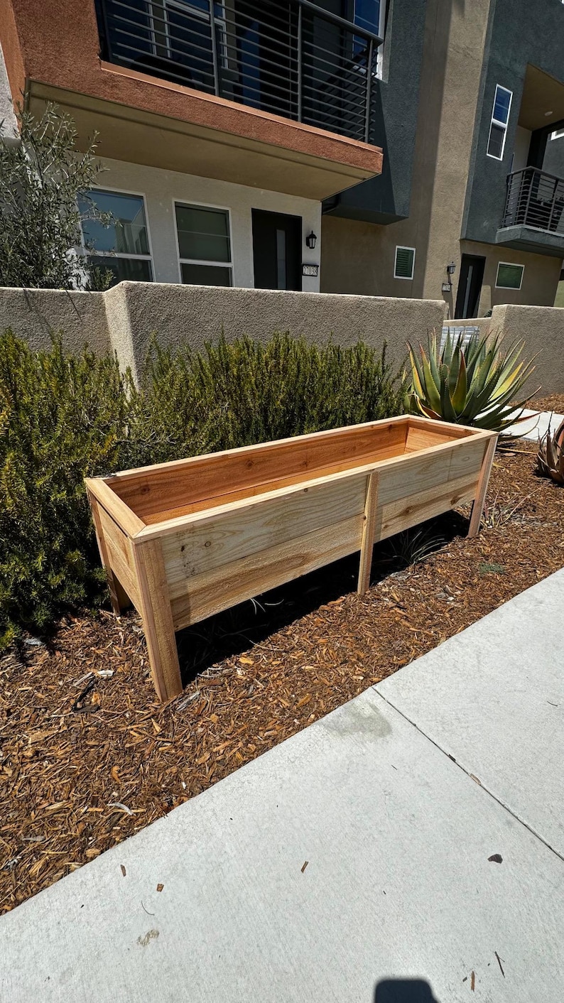 Puede incluir: Una jardinera rectangular de madera con un acabado natural en color marr&oacute;n claro, que cuenta con cuatro patas resistentes y un interior profundo y abierto. La jardinera est&aacute; construida con tablones de madera horizontales y un borde superior reforzado.
