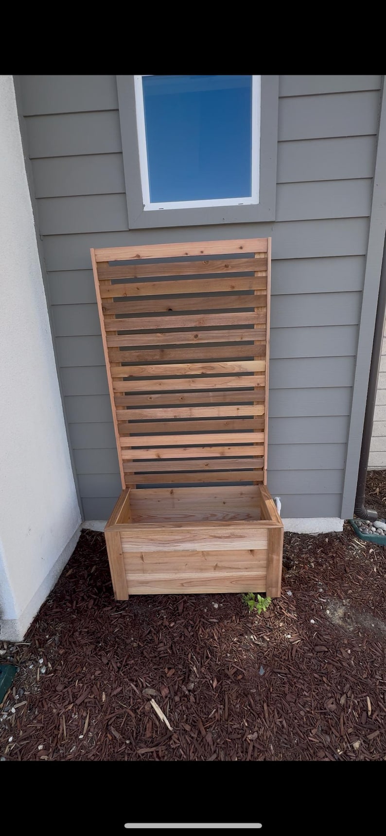 Puede incluir: Una jardinera rectangular de madera con un panel trasero de enrejado vertical alto e integrado. La estructura est&aacute; construida con tablones de madera de color marr&oacute;n claro natural con listones horizontales y una base profunda.