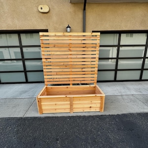 May include: A large, rectangular wooden planter with a slatted trellis back. The planter is made of light-colored wood and sits on a concrete surface. The trellis is open, allowing for climbing plants or decorative elements.