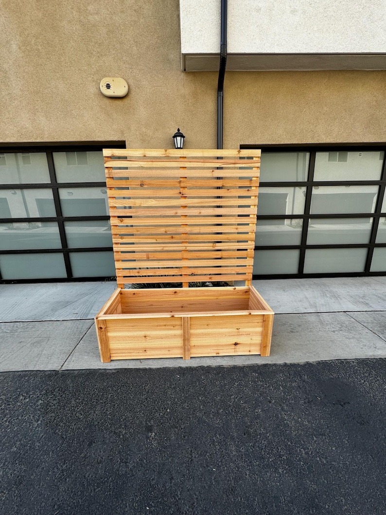 May include: A large, rectangular wooden planter with a slatted trellis back. The planter is made of light-colored wood and sits on a concrete surface. The trellis is open, allowing for climbing plants or decorative elements.